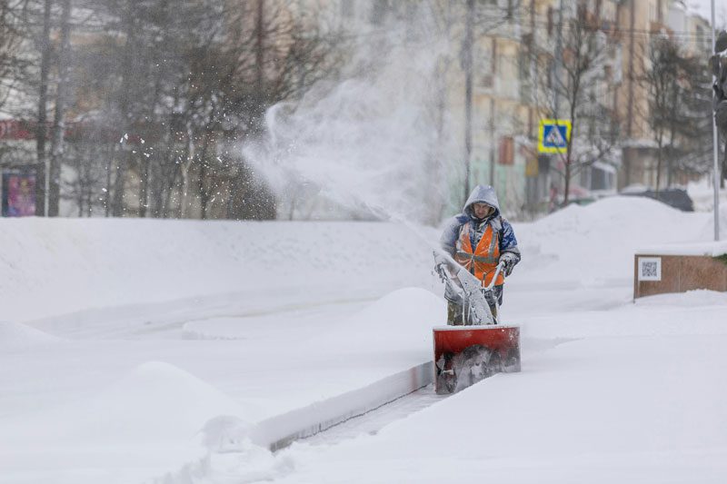 man pushing a snow blower through snowy sidewalk